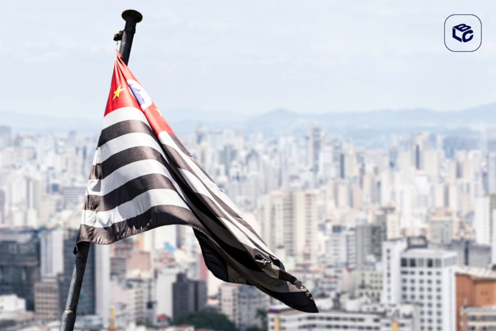 Bandeira do estado de São Paulo tremulando em um mastro, em primeiro plano, com a cidade de São Paulo desfocada ao fundo. A bandeira apresenta listras horizontais pretas e brancas e um canto vermelho com um mapa do Brasil e estrelas. Céu claro e paisagem urbana com prédios altos ao fundo.