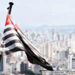 Bandeira do estado de São Paulo tremulando em um mastro, em primeiro plano, com a cidade de São Paulo desfocada ao fundo. A bandeira apresenta listras horizontais pretas e brancas e um canto vermelho com um mapa do Brasil e estrelas. Céu claro e paisagem urbana com prédios altos ao fundo.